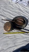 Overhead photo showing three copper pans with long handles arranged on a white surface with measuring tape nearby. Pans show tarnish, discoloration, and surface corrosion.