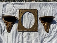 Overall view of two gold vintage wall shelves and one oval picture frame arranged on white cloth outdoors, showing the design and condition of all items.