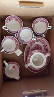 Box containing four pink transferware tea cups, sugar container with cracked handle, and various plates with floral pink designs.