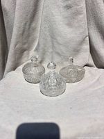 Three clear crystal covered candy dishes with different cut patterns and lid knob shapes on white fabric background.