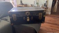 Front angled view of vintage crate trunk showing black metal body, brass corner protectors, rivet detail, and lock clasp without key.