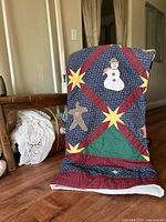 Quilt standing upright showing Christmas appliqués including a snowman, gingerbread man, stars, and a Christmas tree with a plaid border.