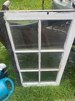 Antique white-painted wooden window with six intact glass panes resting on grass outdoors.