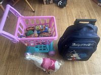 Toy shopping cart with groceries, Mickey Mouse travel bag, and white dancing horse toy arranged on wood floor.