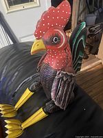 Frontal and side views of the brightly painted wooden rooster figurine showing detailed paintwork on the comb, beak, and wings in red, yellow, black, white, and purple tones.