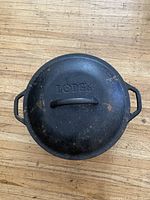 Top view of the cast iron Lodge Dutch oven lid showing Lodge branded embossed on top, placed on wood floor.
