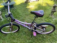Side view of purple girls bicycle with 20 inch wheels on grass, showing frame, wheels, pedals, and seat