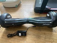 Black Jetson hoverboard with charger visible next to it, showing branding, wheel tread, and charging port side.