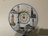 Front view of lighthouse wall clock showing six lighthouse images and seashell decorations around central clock face with gold numbers and black hands.