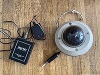 Top view of dome dummy security camera with attached cables, Holland audio simulator box, and transformer plug on wooden surface.