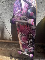 Photo of skateboard showing pink and purple graphic side with guitar and 'Christine' text, purple wheels including one shiny rear wheel, and metallic trucks.
