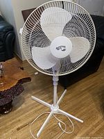 Full view of the white Hampton Bay tall pedestal fan showing fan head, blades, protective wire grille, and tripod base stand.