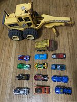 Top view of 1 yellow backhoe loader toy vehicle, 1 yellow dump truck toy vehicle, and 12 various small toy cars in different colors and designs on wooden background.