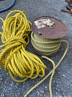 Two rolls of rope on pavement, one yellow synthetic loosely coiled, one beige natural fiber on metal spool showing wear.
