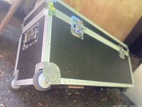 Photo of black wheeled case showing one wheel and metal corner guard on a textured surface outdoors.