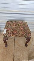 Front view of the embroidered foot stool showing the floral upholstery and carved wooden legs.