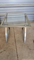 Front angled view of a rectangular metal end table with a clear glass top and curved metal legs, placed on a concrete floor.