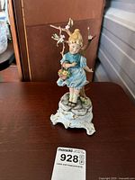 Full front view of porcelain figurine showing girl in blue dress leaning on flowering tree branch, with ornate white and gold base.