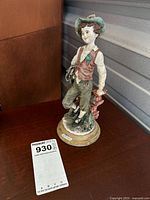 Photo of the full figurine showing boy wearing green hat, brown vest, white shirt, green pants, standing on floral base, leaning on brick pillar.