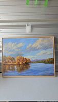 Full image of the framed acrylic landscape painting showing a river or lake with autumn trees, hills in the background, and a bright sky with clouds.