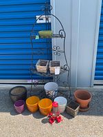 Photo showing black metal corner plant stand with various ceramic and clay pots arranged in front including colorful and neutral tones, plus a red hummingbird feeder.