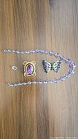 Photo showing purple faceted bead necklace, butterfly ornamental pin with multicolored stones, square pin with large oval purple stone, and small silver ring against wooden surface.