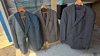 Three men's suit jackets hanging side by side on wooden hangers: dark navy, brown houndstooth, and dark gray pinstripe.