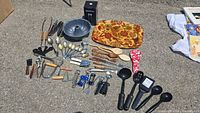 Full view of assorted kitchen utensils including spoons, ladles, knives, corkscrews, decorative ceramic dish, and bundt pan on pavement.