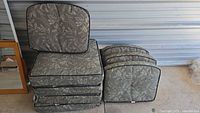 Four outdoor patio chair cushions stacked and arranged against a metal wall. Two back cushions visible standing upright and two seat cushions stacked horizontally, showing grey leaf pattern fabric and black piping.