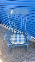 Full front view of blue wooden rocking chair with vertical spindles and a floral stencil on the top backrest. The chair has a blue and yellow plaid padded seat cushion.