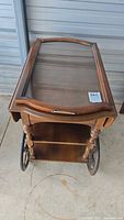 Front view of wooden tea cart showing glass top, wooden frame, metal handle, drawer, two shelves, and large spoked wheels on each side.