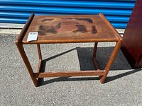 Front and top view of a rectangular teak wood end table with visible stains and wear marks on the tabletop, photographed outdoors in sunlight.