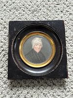 Full view of the framed miniature portrait painting showing the black square wooden frame and the circular gold-tone bezel framing the portrait.