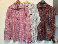 Three men's dress shirts hanging on wall, showing colors and patterns: pink with black geometric pattern, white with black irregular lines, multicolor abstract pattern.