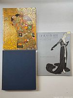 Three hardcover books laid flat: Klimt book with golden ornate cover, Erté fashion illustration book with gray cover, and a plain dark blue hardcover book.