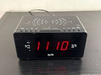 Front view of black SXE alarm clock showing red LED display time 11:10, labels for alarm and PM indicator, and wireless charging symbol on front.