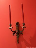 Front view of antique brass two-candle wall sconce mounted on a red wall, showing tall red tapered candles and detailed acorn and foliage design.