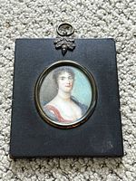 Front shot of rectangular black wooden frame with oval miniature portrait of a woman, showing the overall artwork and frame with metal hanging detail at top.