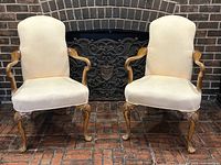 Front view of the pair of beige upholstered antique French armchairs with carved wooden arms and cabriole legs, showing fabric stains.