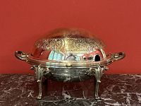 Front view of the antique silver domed revolving chafing dish with ornate floral embossing on the lid and curved legs.