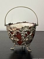 Front view of an oval silver plated sugar basket with oak leaf and acorn decoration, maroon stoneware liner, and three legs, one slightly bent.