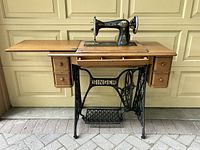 Front view of antique Singer sewing machine mounted on wood table with drawers and cast iron foot pedal base.