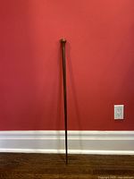Full length view of antique wooden walking stick standing against red wall showing overall shape, handle, and tip.