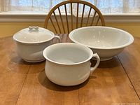 Photo showing the ironstone jug, bowl, and chamber pot with lid on a wooden table with a chair and window in the background.
