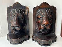 Front view of two carved wood bookends each featuring a lion’s head with worn dark finish.