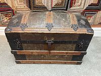 Front view of the antique dome top steamer trunk showing wooden panels, metal reinforcements, central lock, and weathered surface.