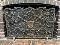 Full front view of wrought iron fire screen positioned in fireplace opening showing full scrollwork detail and brass crest.