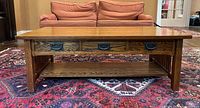 Front view of vintage oak coffee table showing three drawers with black cast metal handles, slatted side panels, and a lower shelf. Sitting on a red patterned carpet with a pink sofa in background.