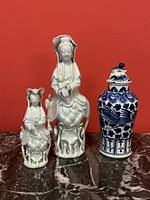 Front view of three Asian porcelain pieces on marble surface against red background. Largest white figurine shows breakage on base.