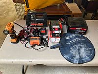 View of all items on white table showing various battery chargers, jumper cables in case, tire inflator, and battery tester.